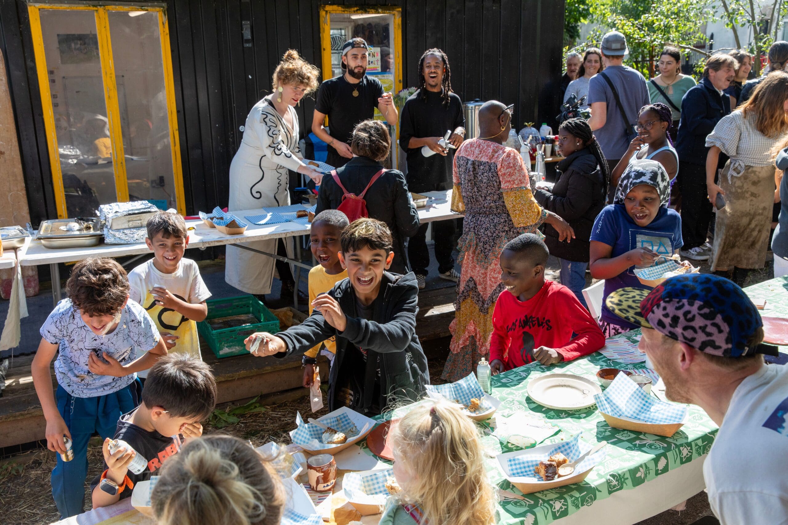 Children around a table laughing and eating with adults in the background serving food